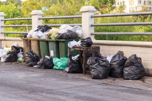 A collection of black, green, and transparent plastic rubbish bags filled with waste is gathered along the edge of an urban street pavement, positioned next to a metal railing and a concrete barrier. Some bags are stacked on top of a green wheelie bin, which is partially obscured. The rubbish bags appear to contain household or office waste, with a variety of textures ranging from crumpled to smooth, and are arranged in a somewhat disorganized manner. In the background, there are leafy trees and a glimpse of a multi-storey building, indicating a residential or mixed-use area. The scene is well-lit with natural daylight, and the overall setting suggests the need for professional rubbish removal services, such as those offered by Office Clearance Harlesden, to manage waste collection efficiently in the local service area near Harlesden or NW10 postcode.