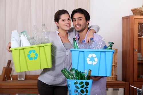 A young couple standing indoors in a room with wooden furniture and cream-colored curtains, each holding a plastic recycling bin filled with empty glass bottles and recyclable plastic containers. The woman, on the left, has dark hair tied back and is wearing a light grey top, while the man, on the right, has dark hair and a beard, wearing a purple shirt. Behind them is a wooden sideboard with additional items, indicating a domestic setting. The bins are brightly coloured, one lime green and the other blue, featuring recycling symbols, and the blue bin has a smaller basket with more glass bottles inside. The scene appears to be part of a rubbish or recycling initiative, supporting proper waste segregation in a household environment, with a focus on recycling glass and plastics commonly collected during rubbish removal services in Harlesden and surrounding London areas.