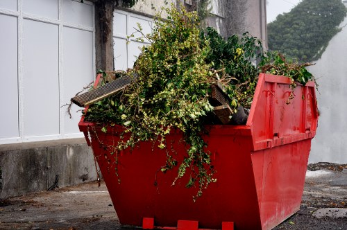 A large red plastic skip filled with green garden waste, including branches, leaves, and small shrubbery, is situated outdoors on a paved area adjacent to a white wooden fence and a concrete wall. The skip, which has a textured surface and a slightly weathered appearance, is positioned on the right side of the image with its lid partially open, revealing the foliage inside. Behind the skip, there is a small tree or bush with dark green leaves, partially visible over the edge of the container. The scene suggests a garden or outdoor clearance, typical of rubbish removal services in London areas like Harlesden. The environment appears clean, well-maintained, and suitable for professional waste collection, highlighting the type of garden rubbish removals provided by Office Clearance Harlesden.