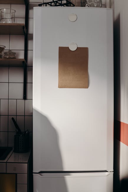 Two women are positioned in a modern kitchen setting. The woman in the background, with long dark hair, is standing and appears to be opening or examining a vintage-style white SMEG refrigerator, whic