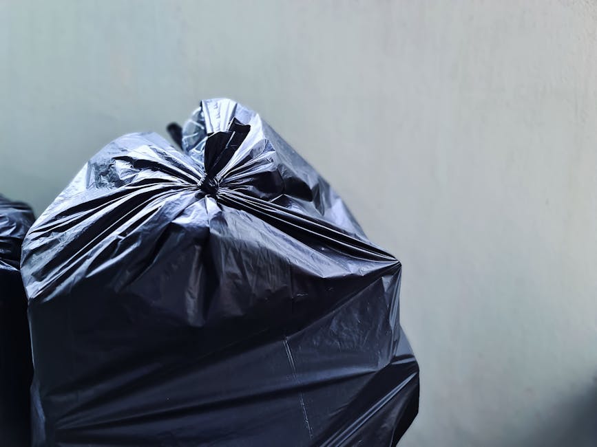 A pile of mixed waste and packaging materials, including flattened cardboard boxes, plastic bags, and paper wrapping, is gathered against a concrete brick wall and a tree trunk outdoors. The cardboard